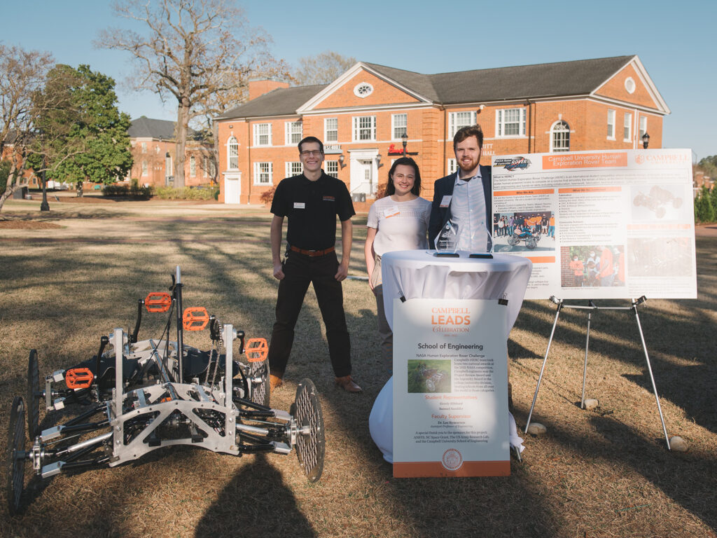 Two young men and a young woman standing beside two posters. One says "Campbell University human exploration rover team" and the other says "Campbell Leads celebration, school of engineering". A four-wheeled human-powered vehicle is on the ground beside them.