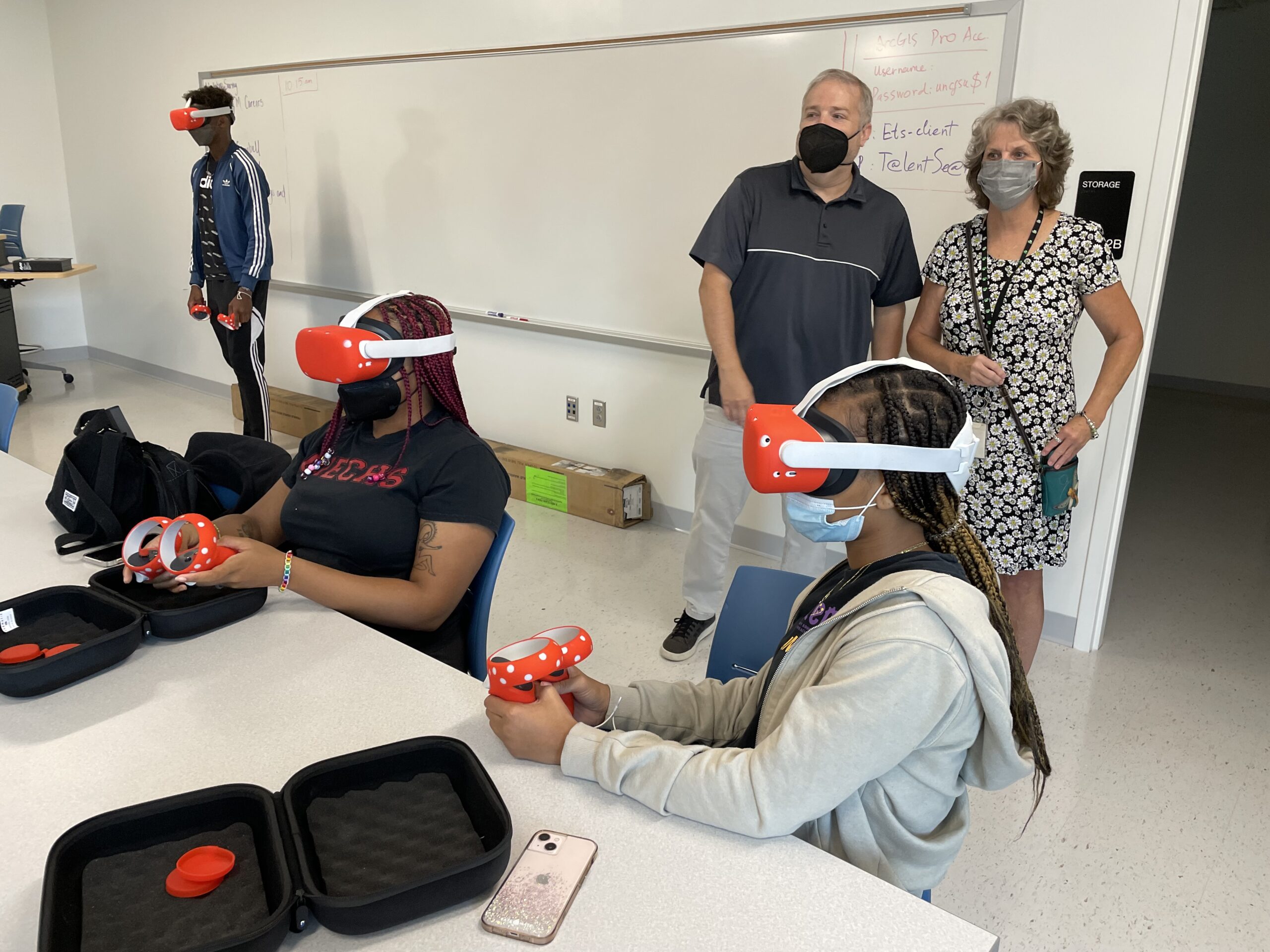 Two students are wearing VR headsets and holding the controllers. Two adults stand in the background supervising them.