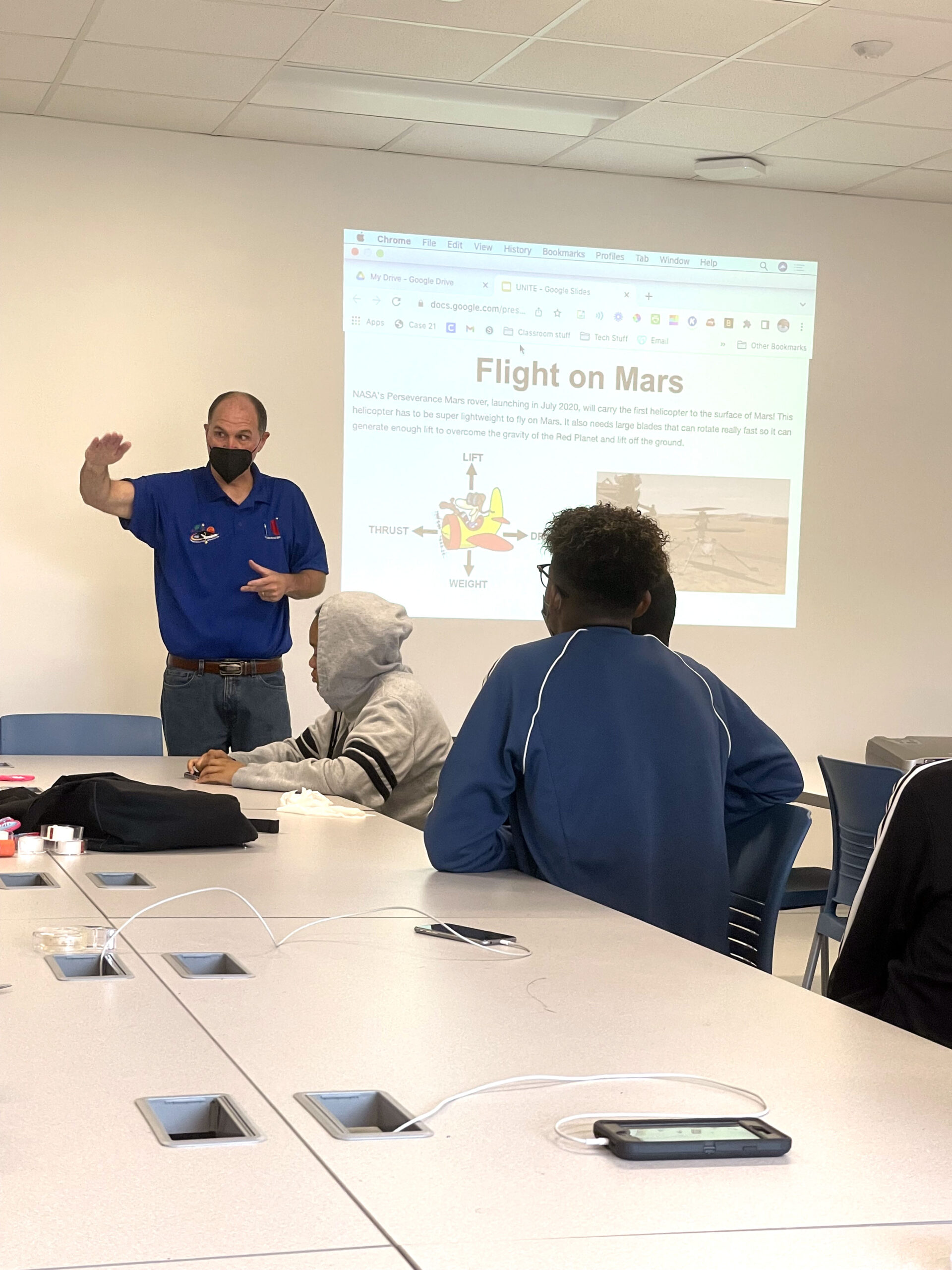 A man stands in front of a projector image titled "Flight on Mars". Three students are sitting at a long desk and listening to the man.