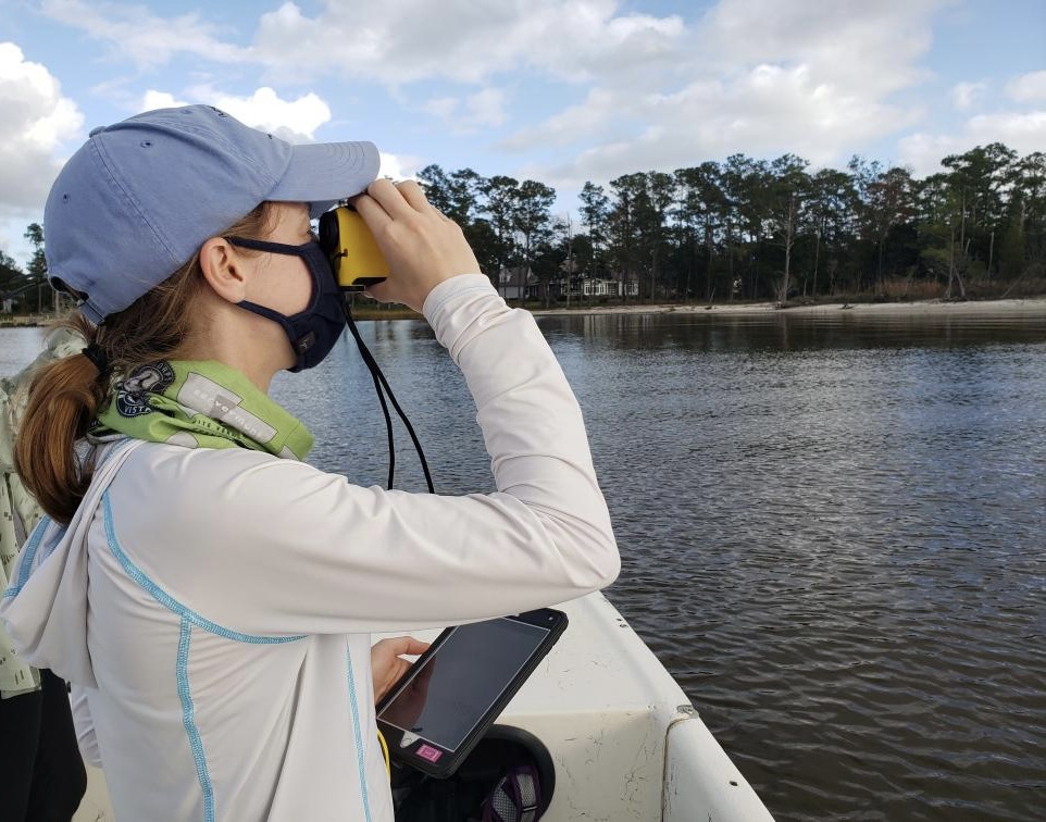 Young woman, looking through binoculars and holding a tablet on a boat near the shoreline