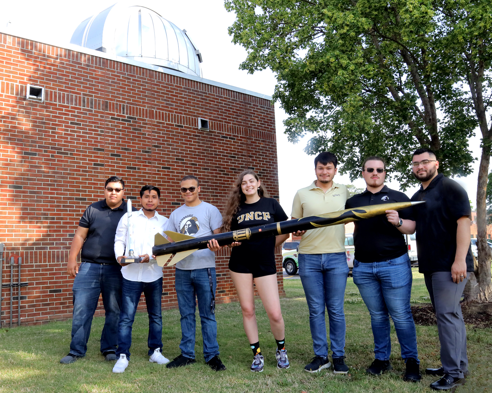 Six young adults are standing in front of a building and carrying a long rocket together.