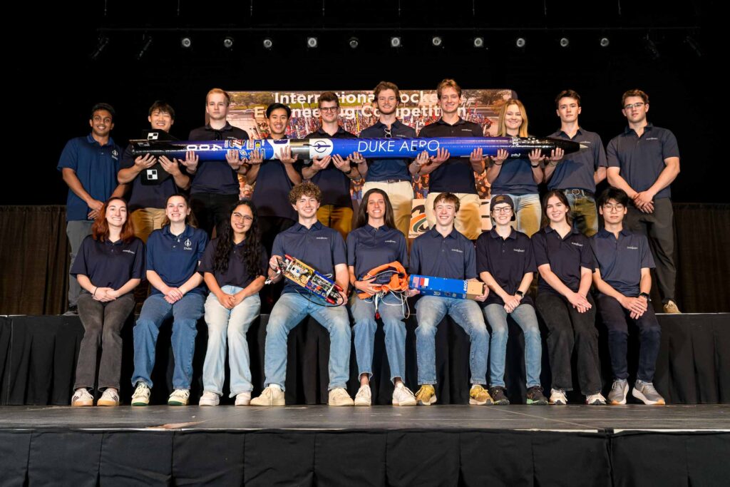 Students in two rows on a stage. The upper row is holding a blue rocket