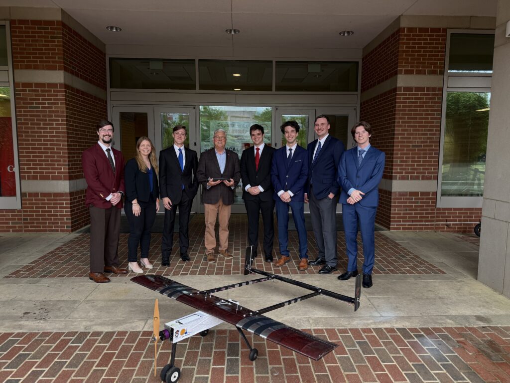 8 people standing in a row near a brick building. A small airplane is in front of the group