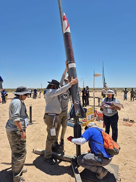 Several people standing in the sand. One is holding up a large rocket while another adjusts the rocket stand.