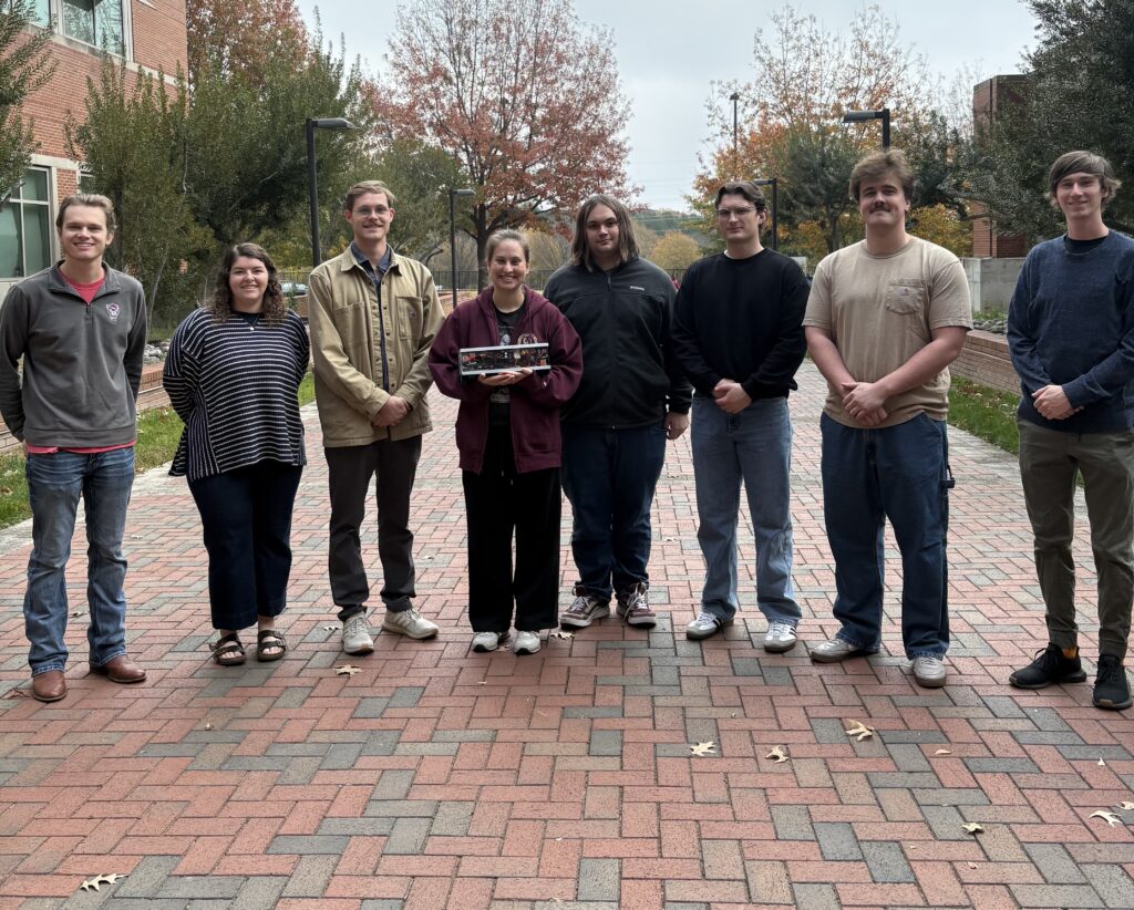 A group of seven people standing next to each other on a brick road holding a small device