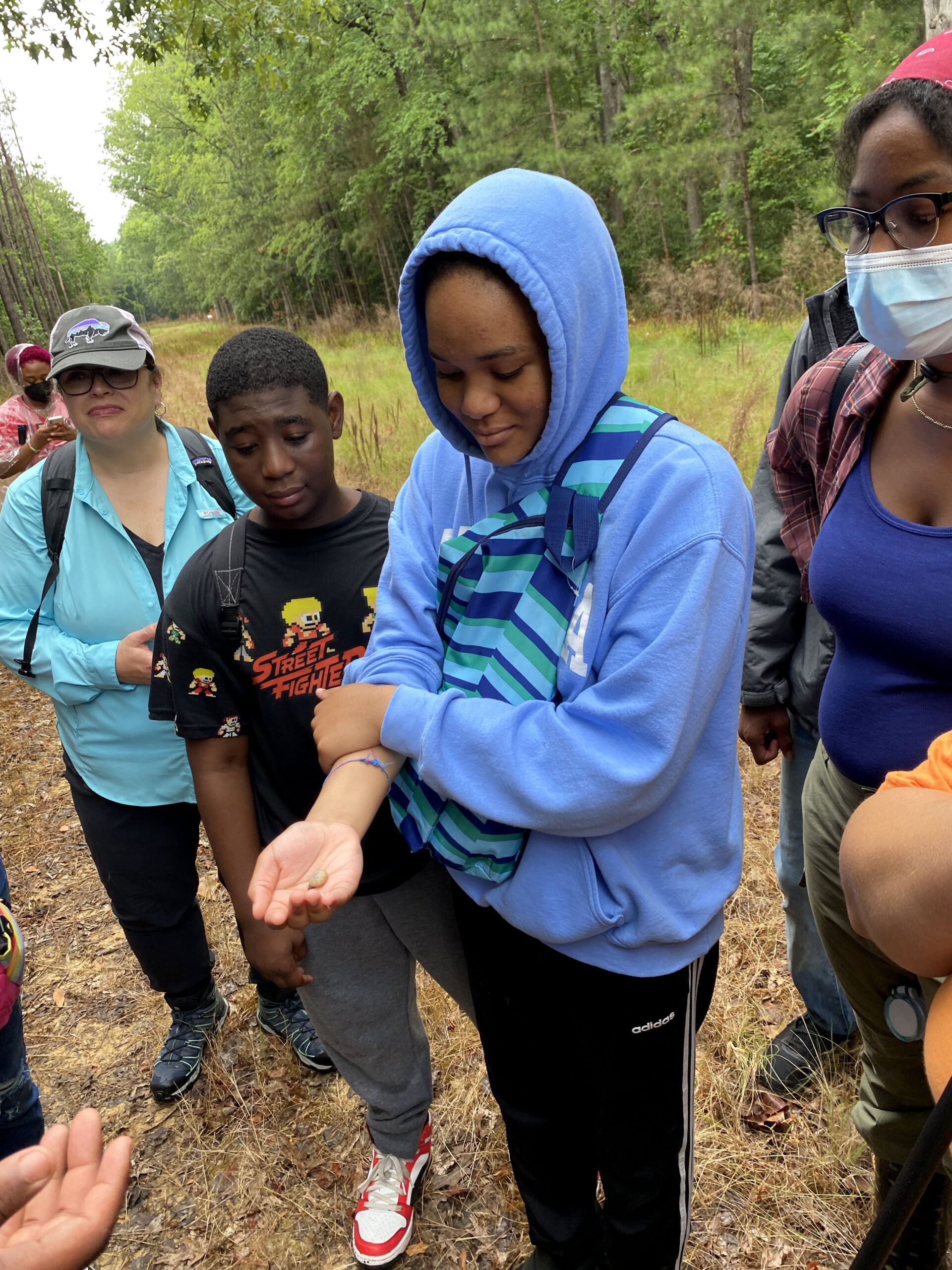 A group of students stand together in a forest clearing, looking at a snail that is curled in one of the students' hand.