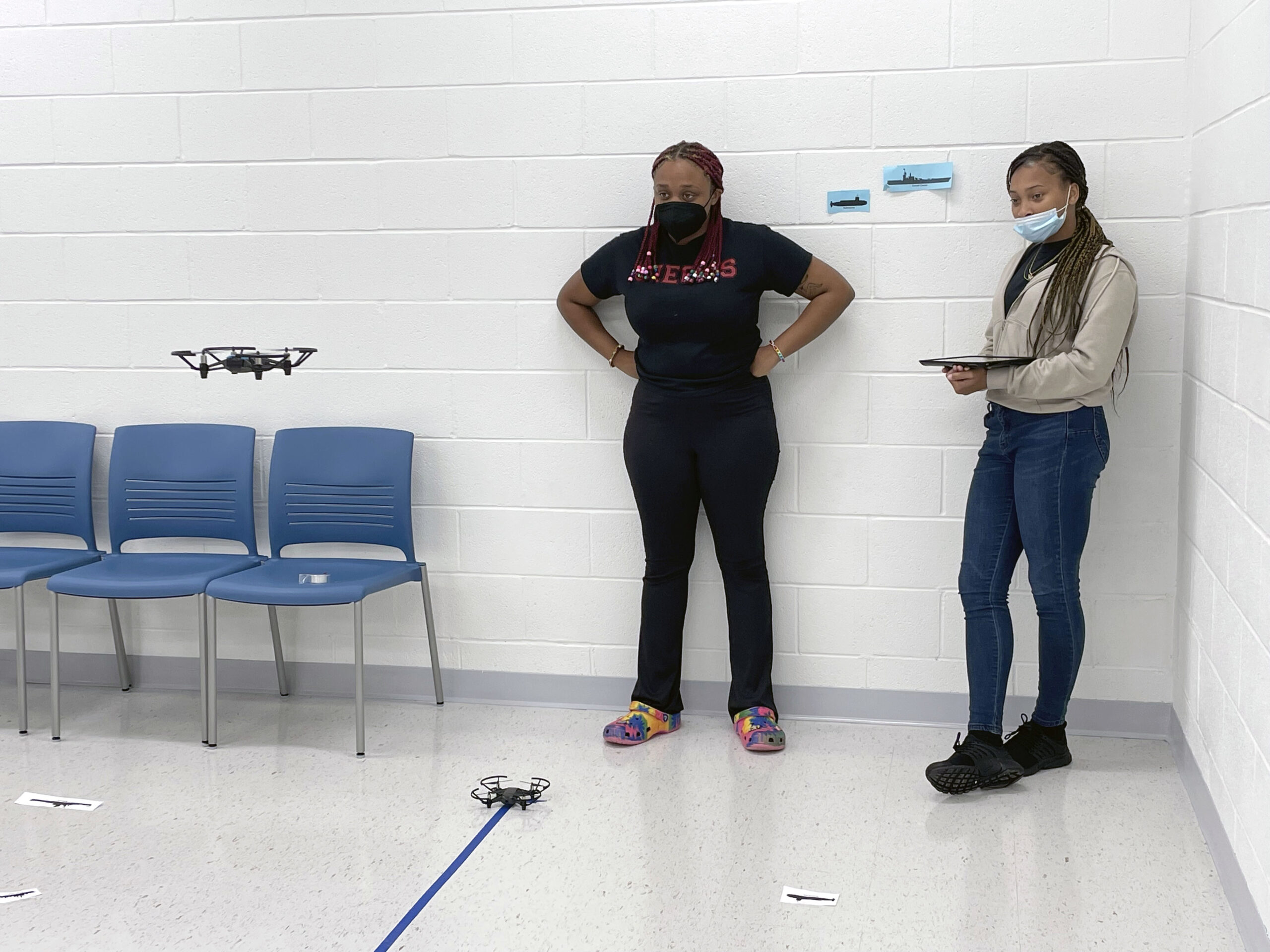 Two young women watching a small remote-controlled aircraft flying in a room. One woman is holding a tablet.