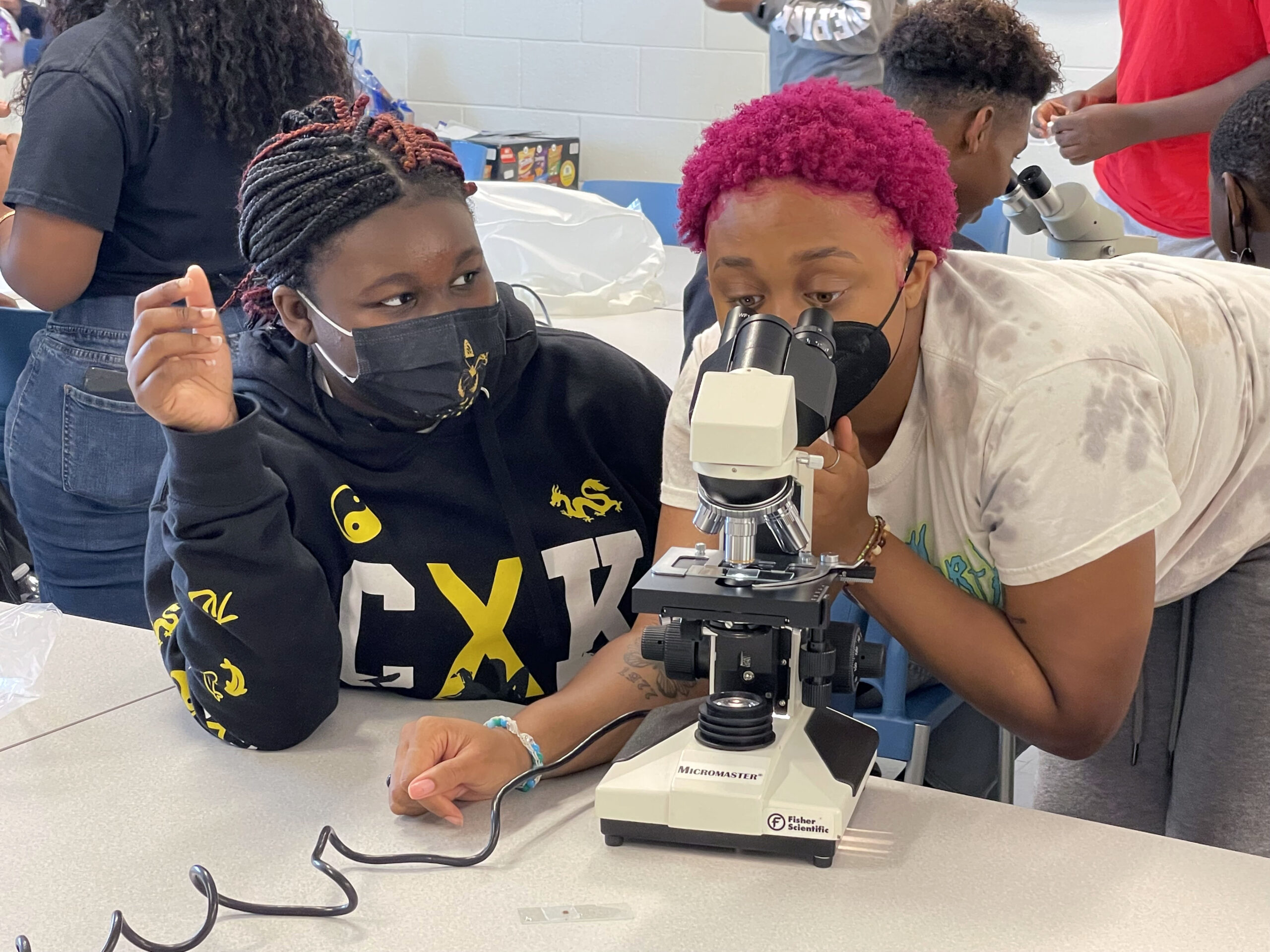 Two students crowd around a microscope. The student on the right is looking through the lens while the student on the right watches on
